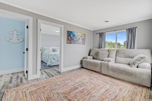 A room with a grey sofa, a colorful rug, a bed with a nautical-themed duvet, and a painting on the wall.