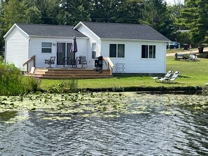A white cabin with a deck and outdoor seating, situated next to a body of water with lily pads.