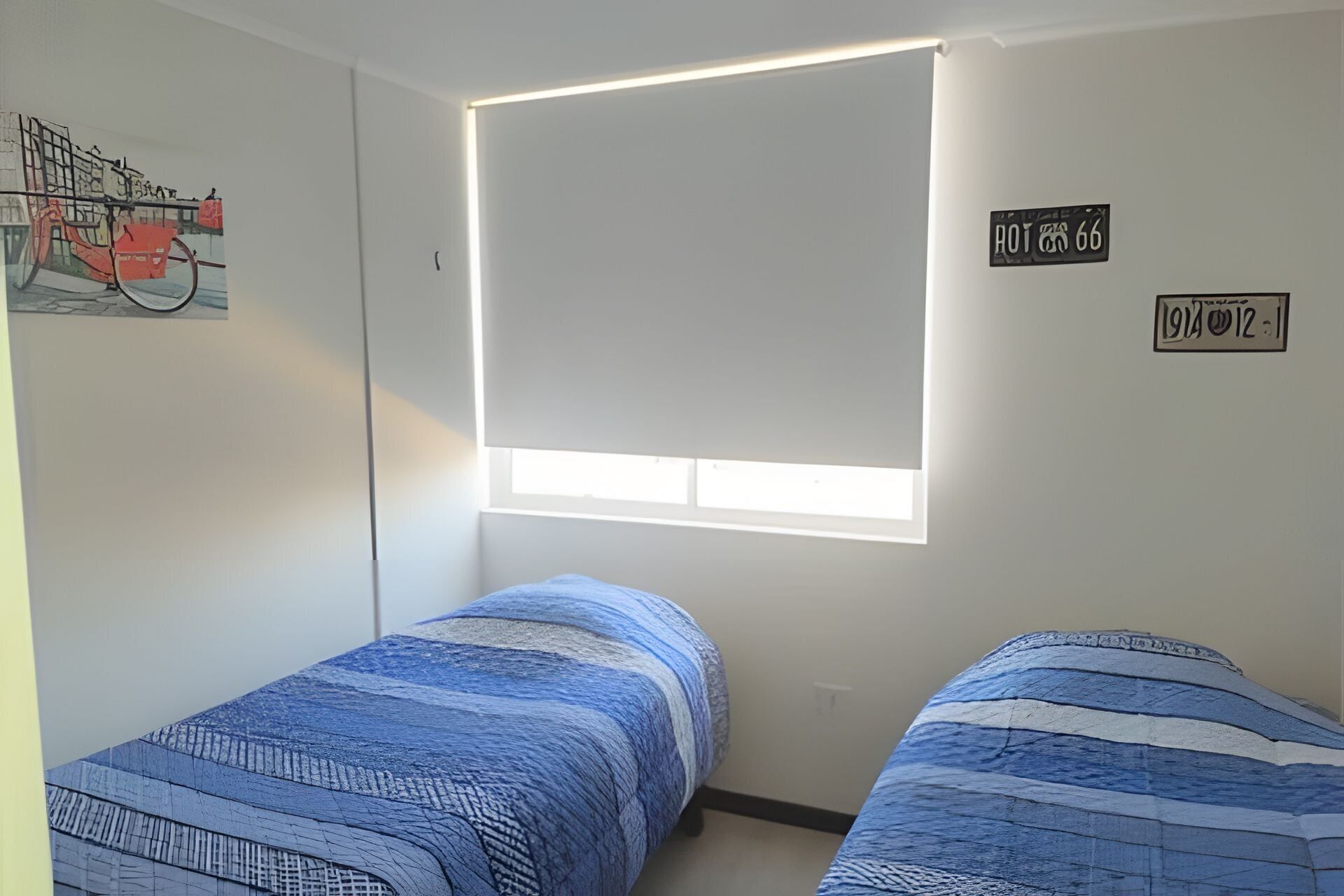 Una habitación con dos camas, una ventana con persianas, una obra de arte colgada en la pared y dos letreros en la pared.