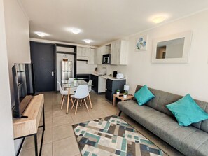 Ein kompakter Wohnbereich mit Küchenzeile, Essplatz und Sofa mit türkisfarbenen Kissen.