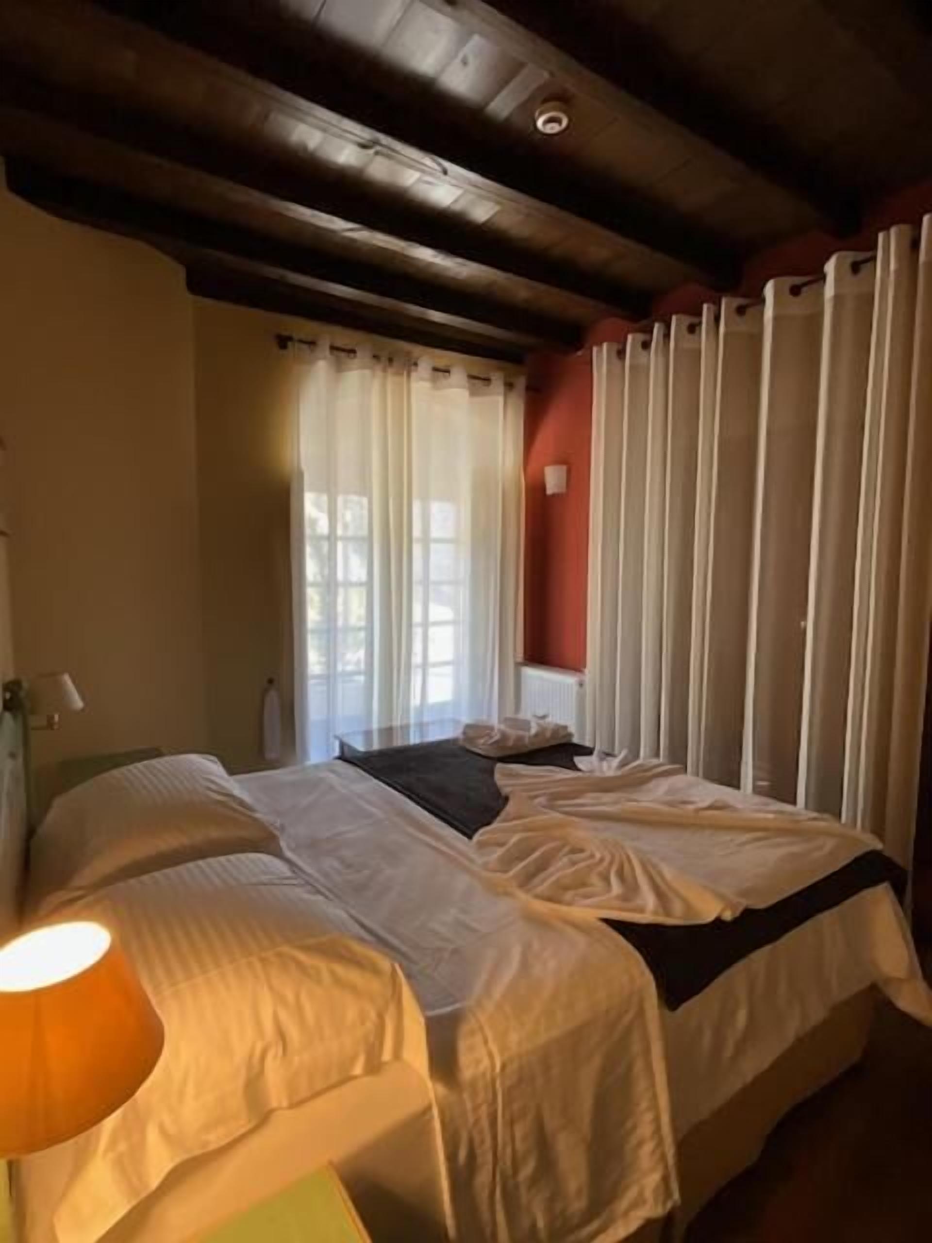 A room with two beds, a wooden ceiling, a window with curtains, and a bedside lamp.