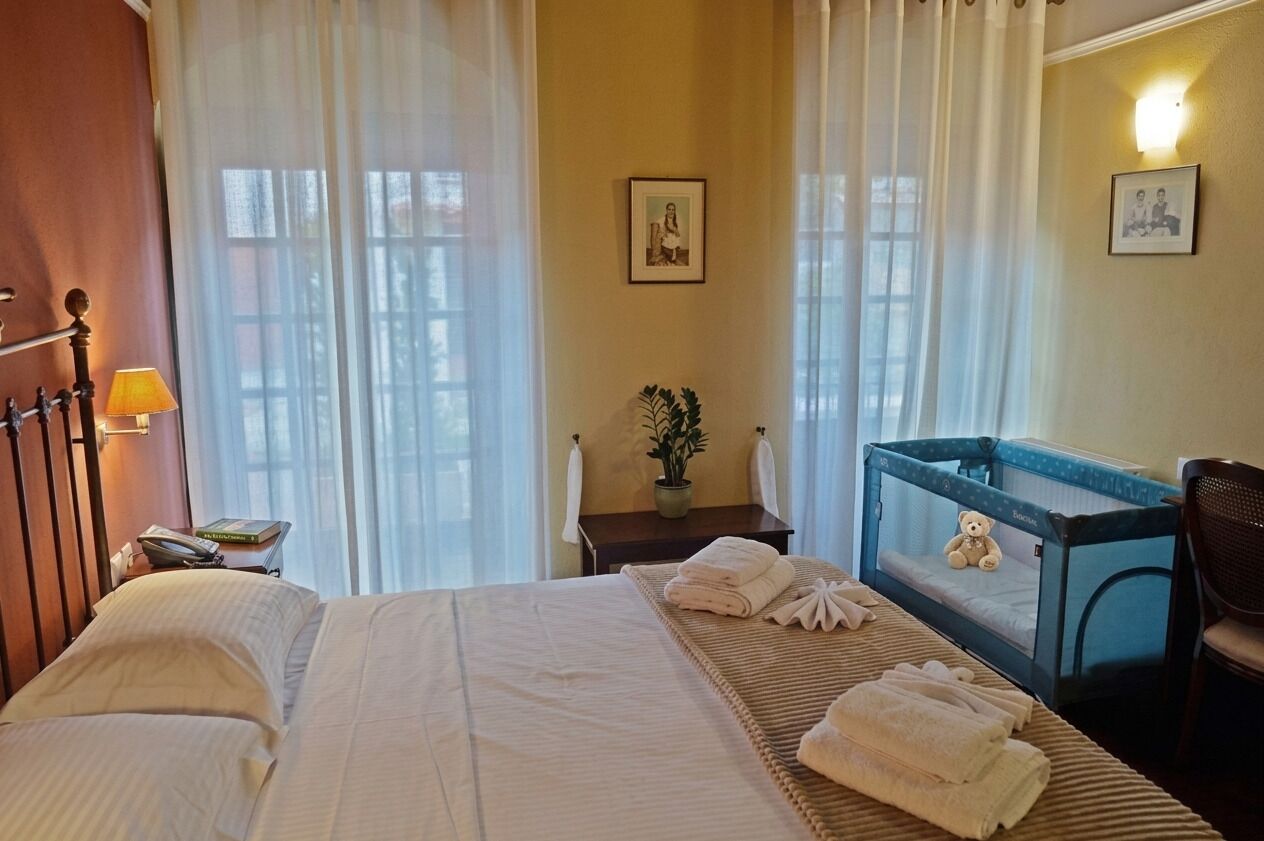 Habitación de hotel con cama, escritorio con lámpara, una silla, cuna con un osito de peluche y planta en maceta.