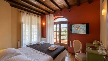 A hotel room with a bed, a desk, a chair, a painting, and a view through an arched window.