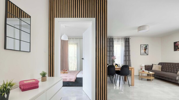 Ein modernes Wohnzimmer mit Sofa, Esstisch und Stühlen. Im Hintergrund steht ein Bett, eine Topfpflanze und eine dekorative Wandverkleidung.