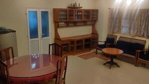 Una sala de estar con una mesa de comedor, sillas, un conjunto de sofá, un aparador con puertas de cristal y una ventana con cortinas.