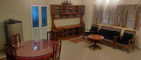 Una sala de estar con una mesa de comedor, sillas, un conjunto de sofá, un aparador con puertas de cristal y una ventana con cortinas.