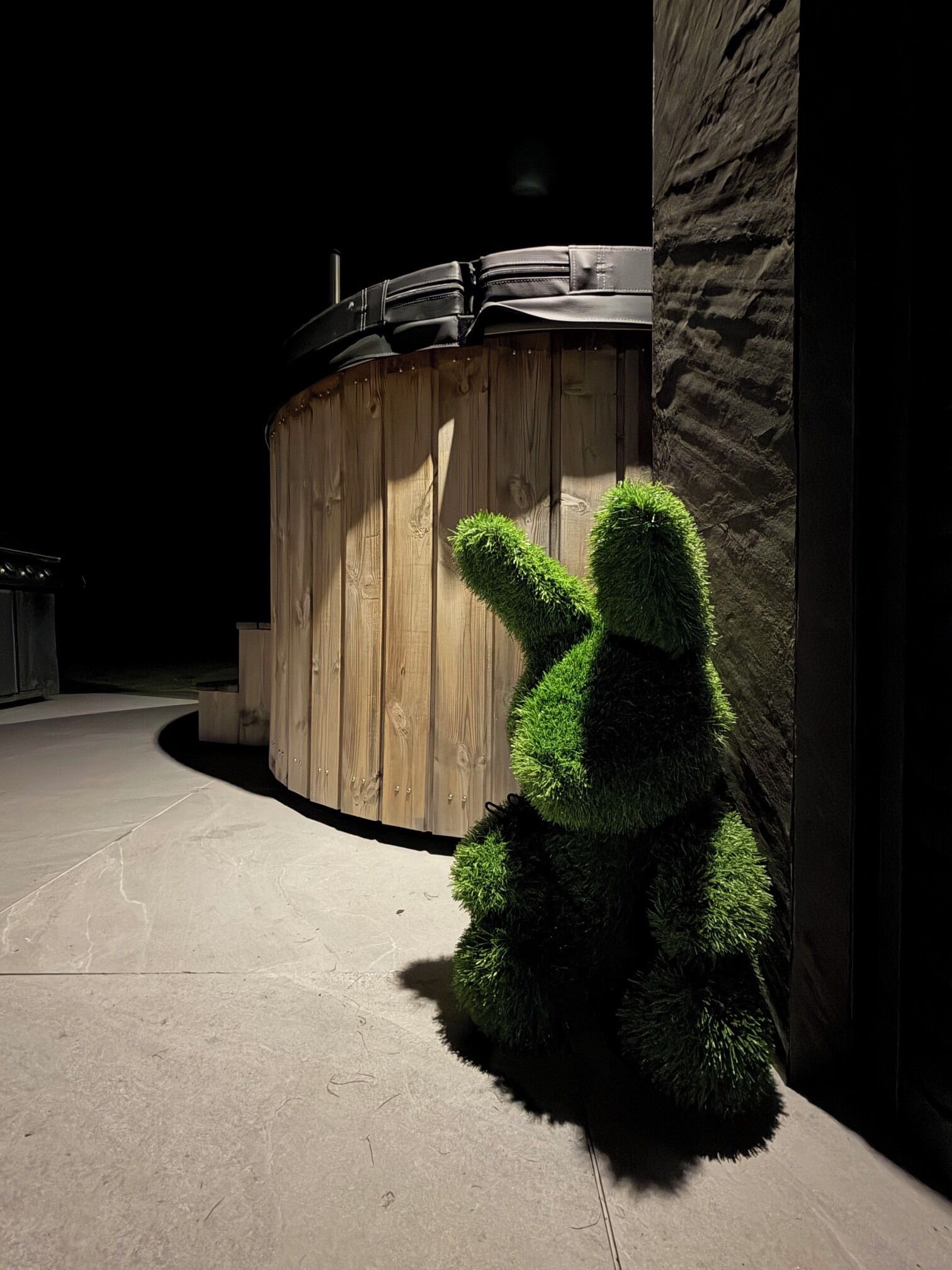 Un jacuzzi con revestimiento de madera, un arbusto artificial verde y una pared de piedra.