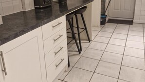 Une cuisine avec des meubles blancs, un plan de travail noir, un grille-pain, une bouilloire et un balai.