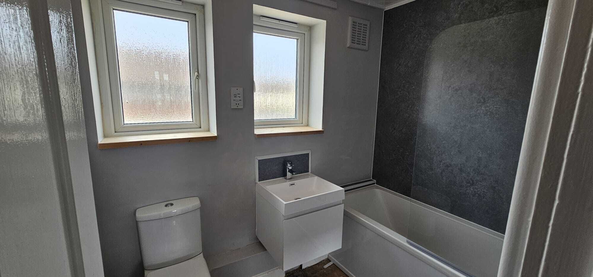 A small bathroom with two windows, a toilet, a sink, and a shower.