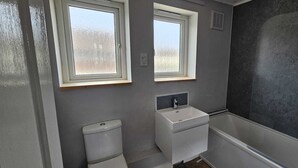 Une petite salle de bain avec deux fenêtres, des toilettes, un lavabo et une douche.