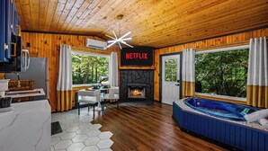Un intérieur de chalet moderne avec une cheminée en pierre, un écran affichant le logo de Netflix, un jacuzzi et un coin repas.