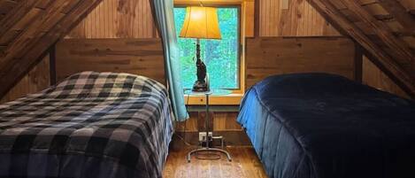 Ett litet sovrum på vinden med träväggar och ett sluttande tak. Det finns två sängar med rutigt sänglinne, en bordslampa och ett fönster med utsikt över grönskan.