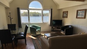 A living room with a large window, a sofa, a television, and a dining table. - Lost Lake Cottage

Munising/Wetmore (Munising Township)