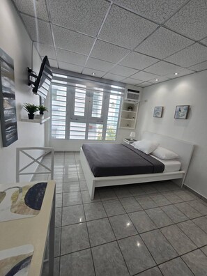 A modern bedroom with a bed, a desk, a chair, a window with blinds, and a ceiling with a grid pattern. - LIS4B12 - SeaSide Beach II (Carolina)