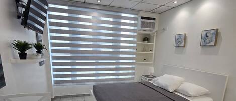 Ein modernes Schlafzimmer mit Bett, Schreibtisch, Stuhl, Fernseher und einem Fenster mit Jalousien.