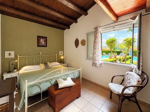 Quarto com cama, mesa de cabeceira, cadeira, janela com cortinas e vista para um jardim.
