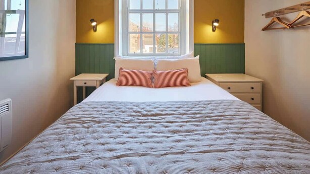 A bed with a striped pillow, two bedside tables, a window with blinds, and a wall-mounted coat rack.