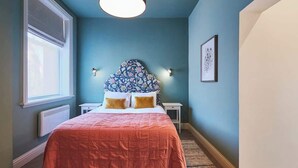 Makuuhuone, jossa on sänky, ikkuna, patteri, seinään kiinnitetty valaisin ja kehystetty taulu.