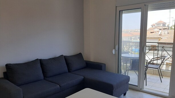 Un canapé de couleur sombre et une table basse blanche se trouvent dans une pièce dotée d’une porte-fenêtre coulissante qui donne sur un balcon extérieur aménagé avec des chaises.