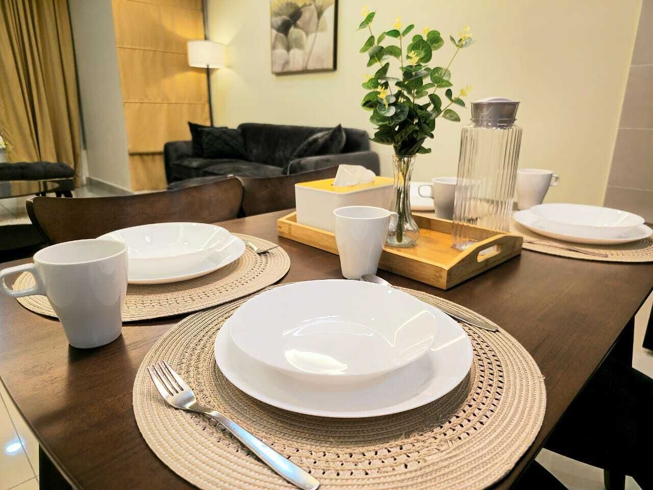 A dining table set with plates, cups, and cutlery, with a vase and a lamp in the background.