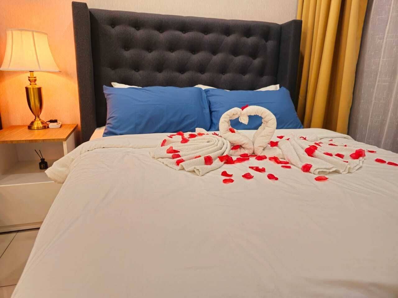 A neatly made bed with a heart-shaped towel arrangement and rose petals, a dark tufted headboard, blue pillows, and a bedside lamp.