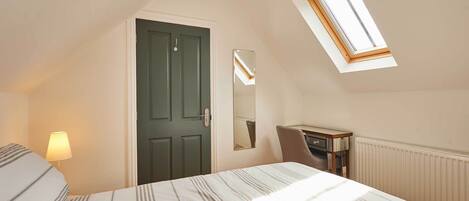 A bedroom with a bed, a desk, a chair, a lamp, a door, and a skylight.