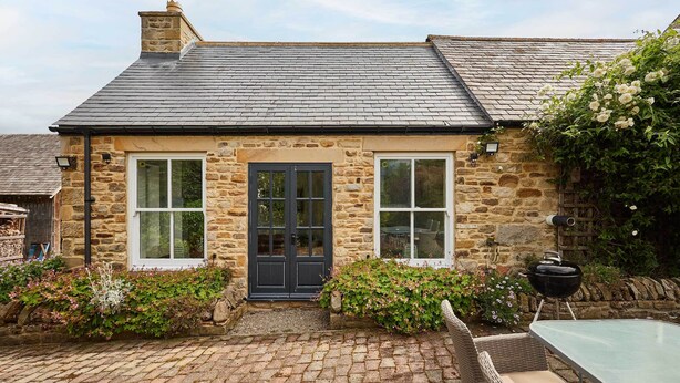 A stone cottage with a dark door, two windows, a brick patio, and a garden with flowers and a grill.