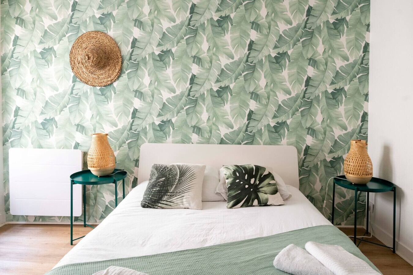 Une chambre à coucher avec un lit, deux tables de chevet, un panier en osier sur chaque table de chevet, un chapeau en osier accroché au mur et un papier peint à motifs de feuilles.