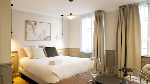 A hotel room with a neatly made bed, a dining table with a wine glass and cutlery, a painting on the wall, and a window with curtains. - Trendy studio apartment, 2 minutes from the very center of town (Caen)