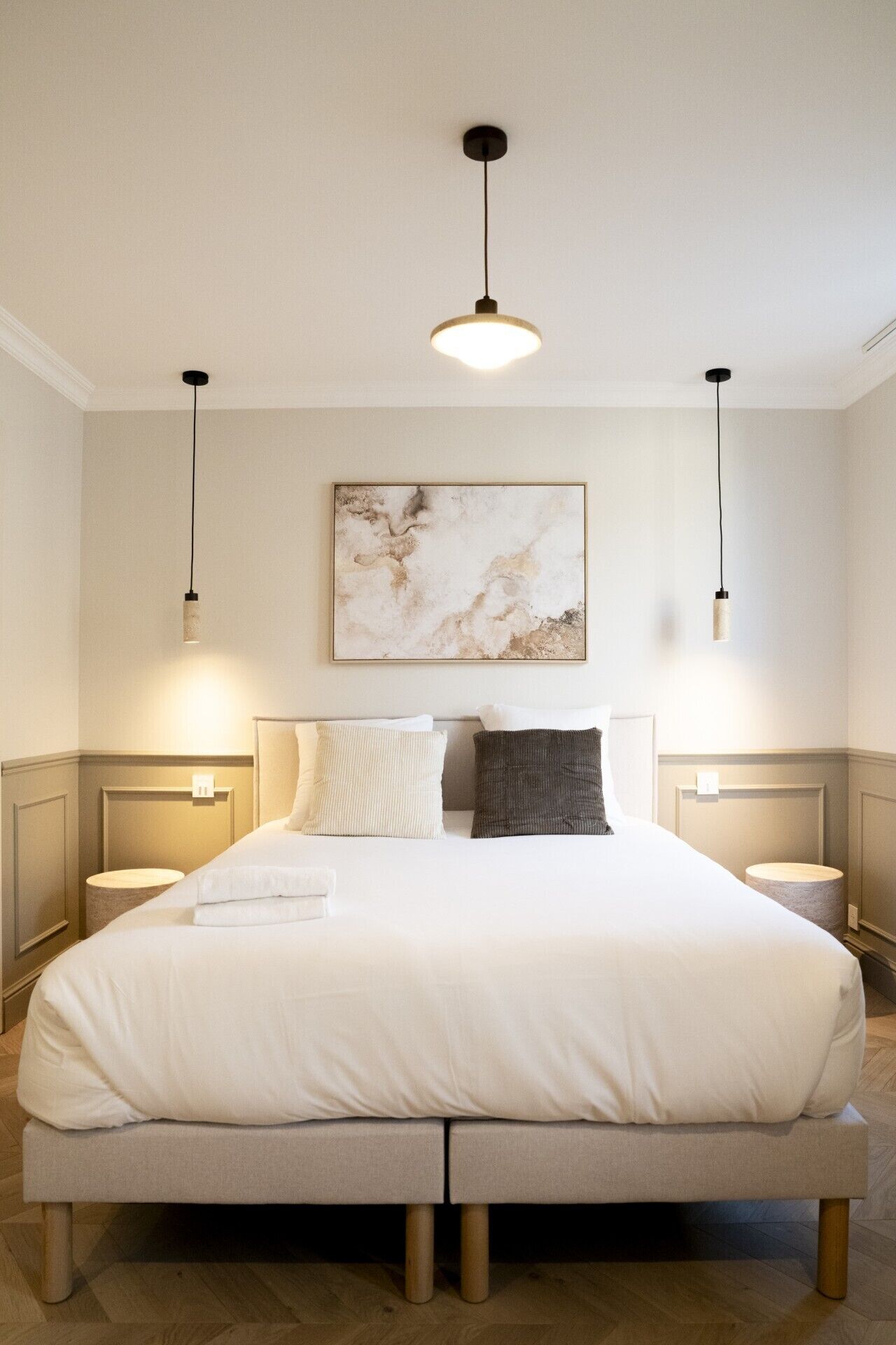 Un grand lit recouvert d’une couette blanche, deux tables de chevet avec des lampes, une œuvre d’art en marbre accrochée au mur et un luminaire suspendu.