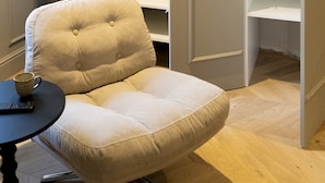 A room with a beige armchair, a small round table with a remote control, and a white shelving unit with open doors. - Trendy studio apartment, 2 minutes from the very center of town (Caen)