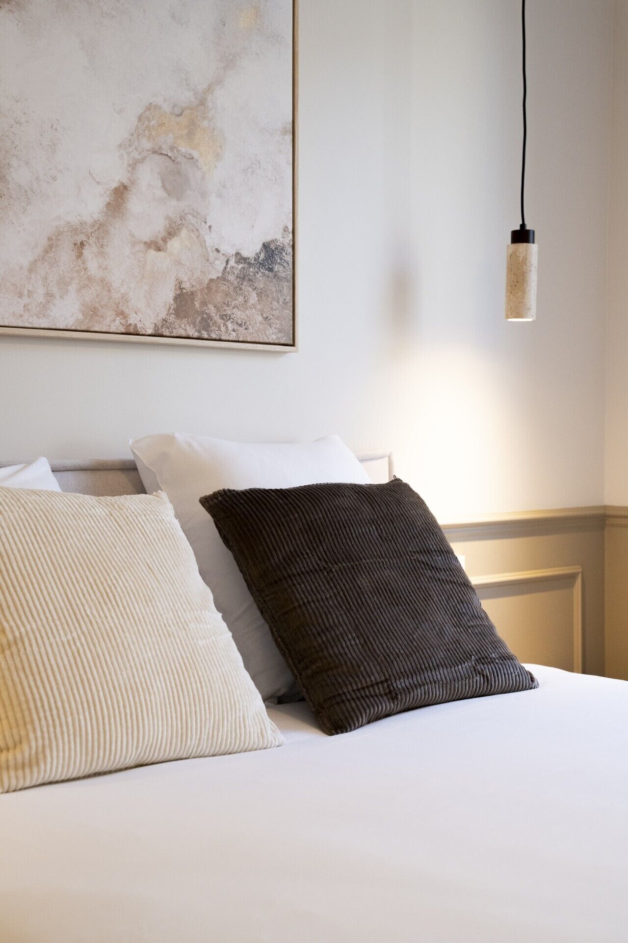 Un lit avec deux oreillers, un mur orné d’un tableau à texture, et une suspension lumineuse.