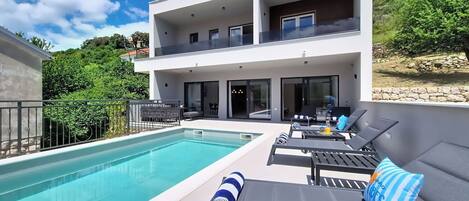Uma casa moderna de dois andares com piscina, área de estar externa e vista para o verde ao redor.