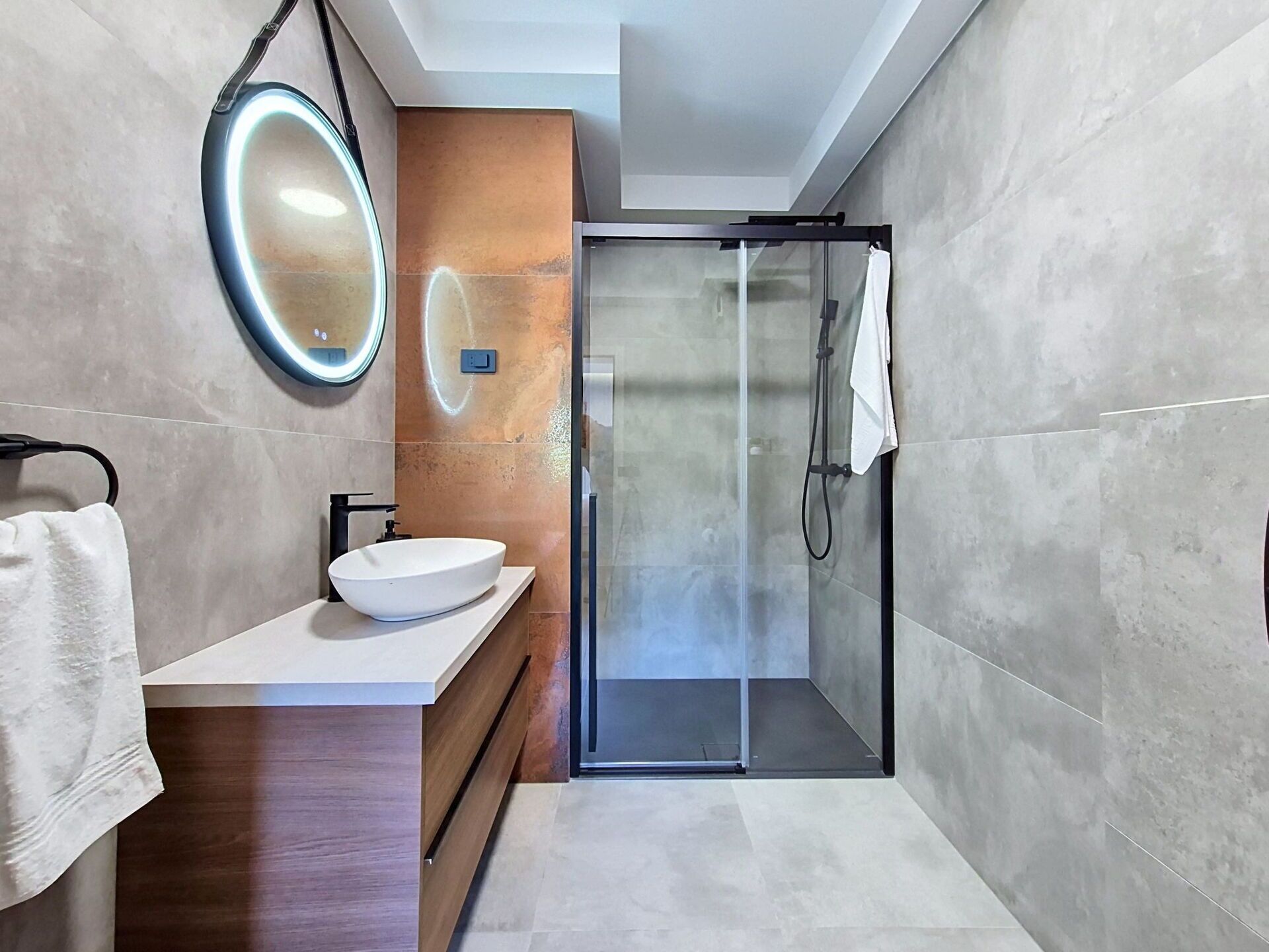A modern bathroom with a glass shower enclosure, a round mirror, and a white sink.