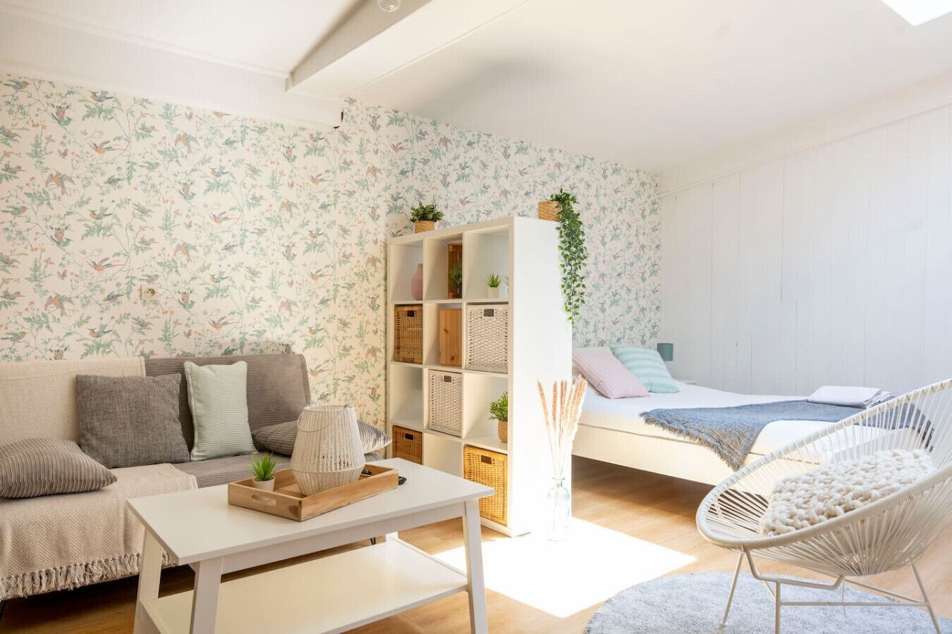 Una sala moderna con un sofá, una mesa de centro blanca, una silla de mimbre y una cama con cabecera blanca, además de una pared con papel tapiz floral.