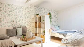 Una sala moderna con un sofá, una mesa de centro blanca, una silla de mimbre y una cama con cabecera blanca, además de una pared con papel tapiz floral.