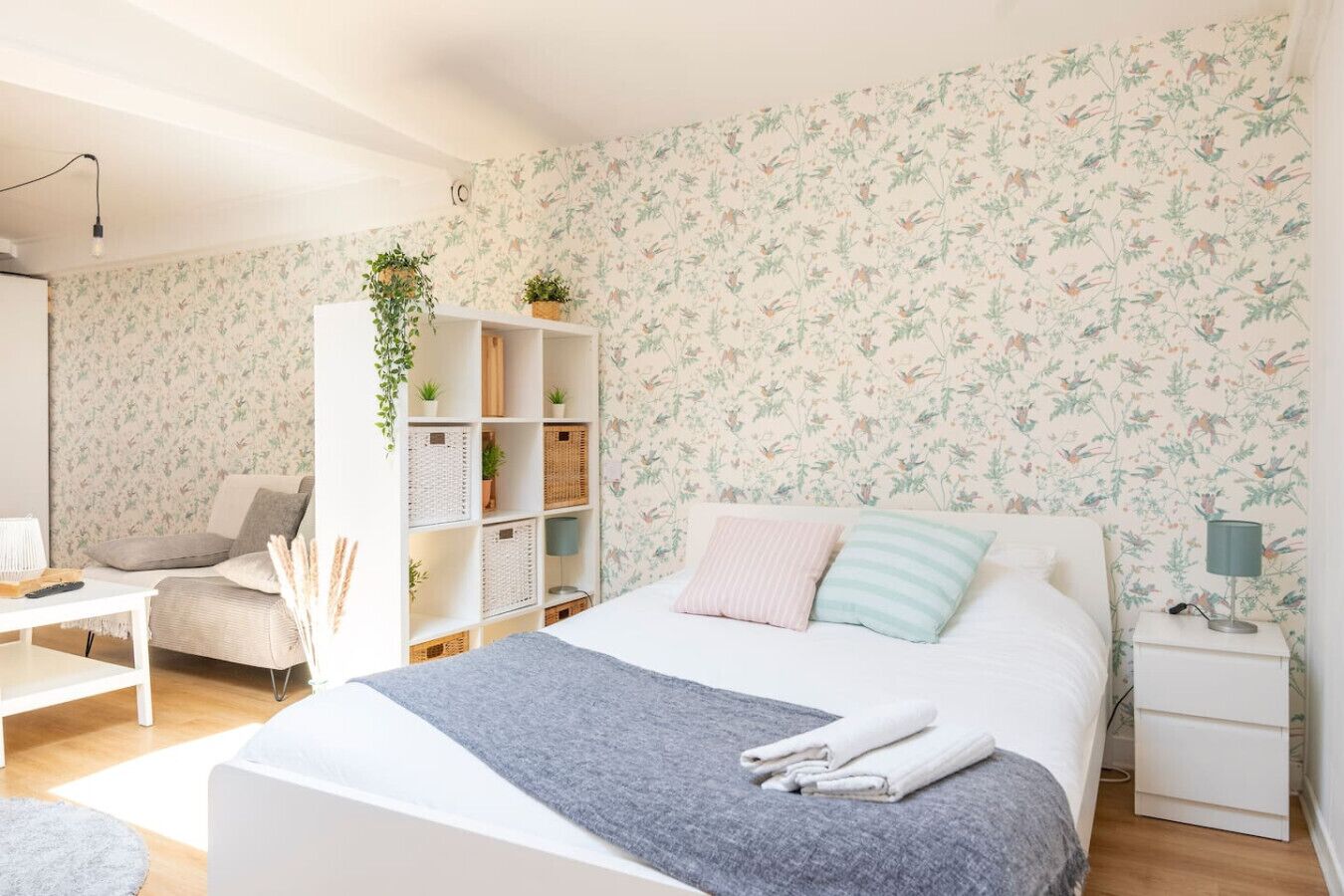 A bedroom with a bed, bedside tables, a desk, a chair, a white wardrobe, and a wall with floral wallpaper.