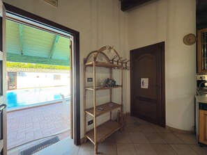 A room with a wooden door, a shelf with items, a clock on the wall, and a view of an outdoor swimming pool. - Wonderful  villa for 5 guests with pool, WIFI, A/C, TV, patio and panoramic view (Chiaramonte Gulfi)