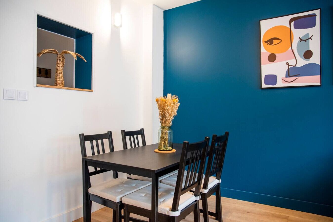 Un coin repas avec une table en bois foncé, quatre chaises, un vase contenant des fleurs séchées, un miroir fixé au mur et une œuvre d’art abstraite.