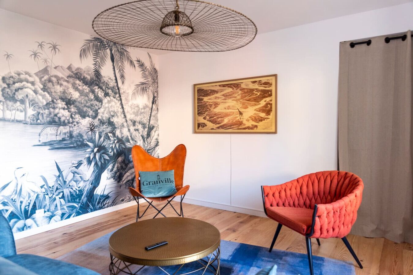 A room with a mural of a tropical scene, a round table, two chairs, a hanging light fixture, and a framed picture on the wall.