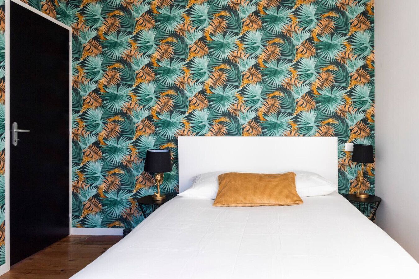Une chambre avec un lit blanc, un coussin décoratif marron, deux lampes de chevet et un mur orné d’un motif de feuilles tropicales.