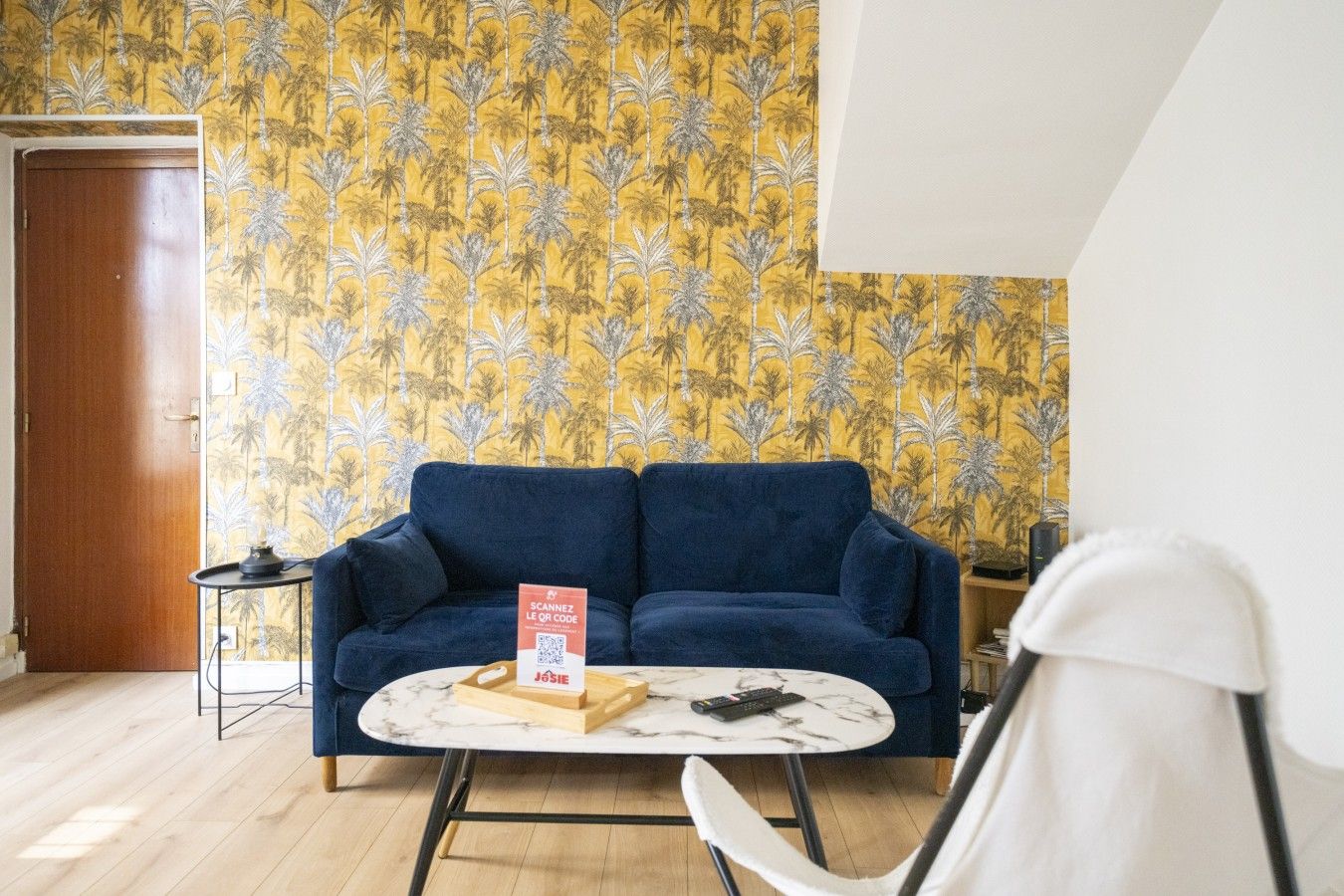 Een kamer met een blauwe bank, een kleine ronde tafel, een witte stoel, een houten bijzettafeltje met een afstandsbediening, en een deur.