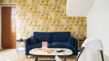 Een kamer met een blauwe bank, een kleine ronde tafel, een witte stoel, een houten bijzettafeltje met een afstandsbediening, en een deur.