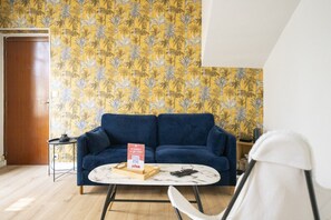 Een kamer met een blauwe bank, een kleine ronde tafel, een witte stoel, een houten bijzettafeltje met een afstandsbediening, en een deur.