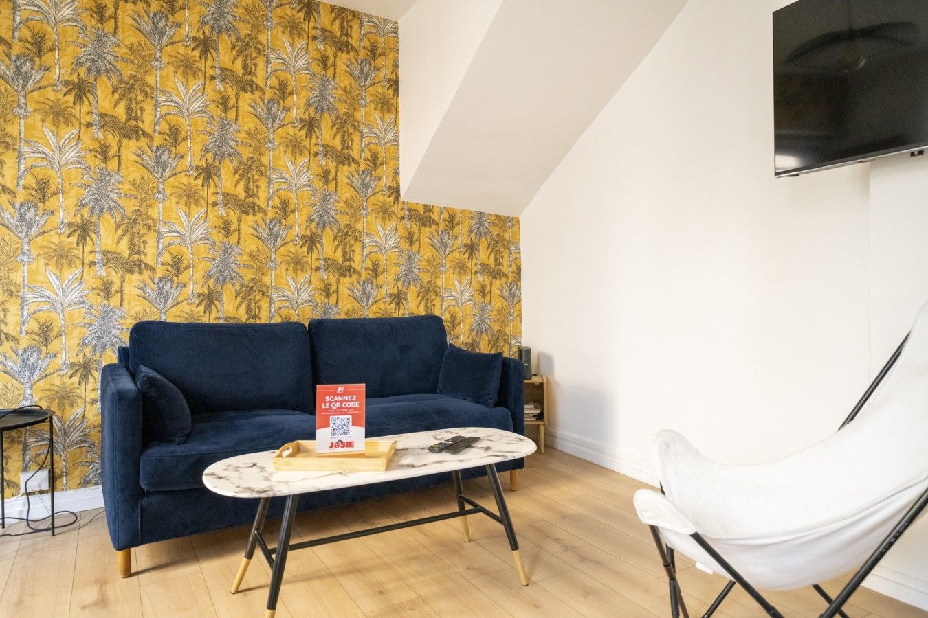 Een kamer met een blauwe bank, een witte stoel, een tafeltje en een muur met een geel patroon.