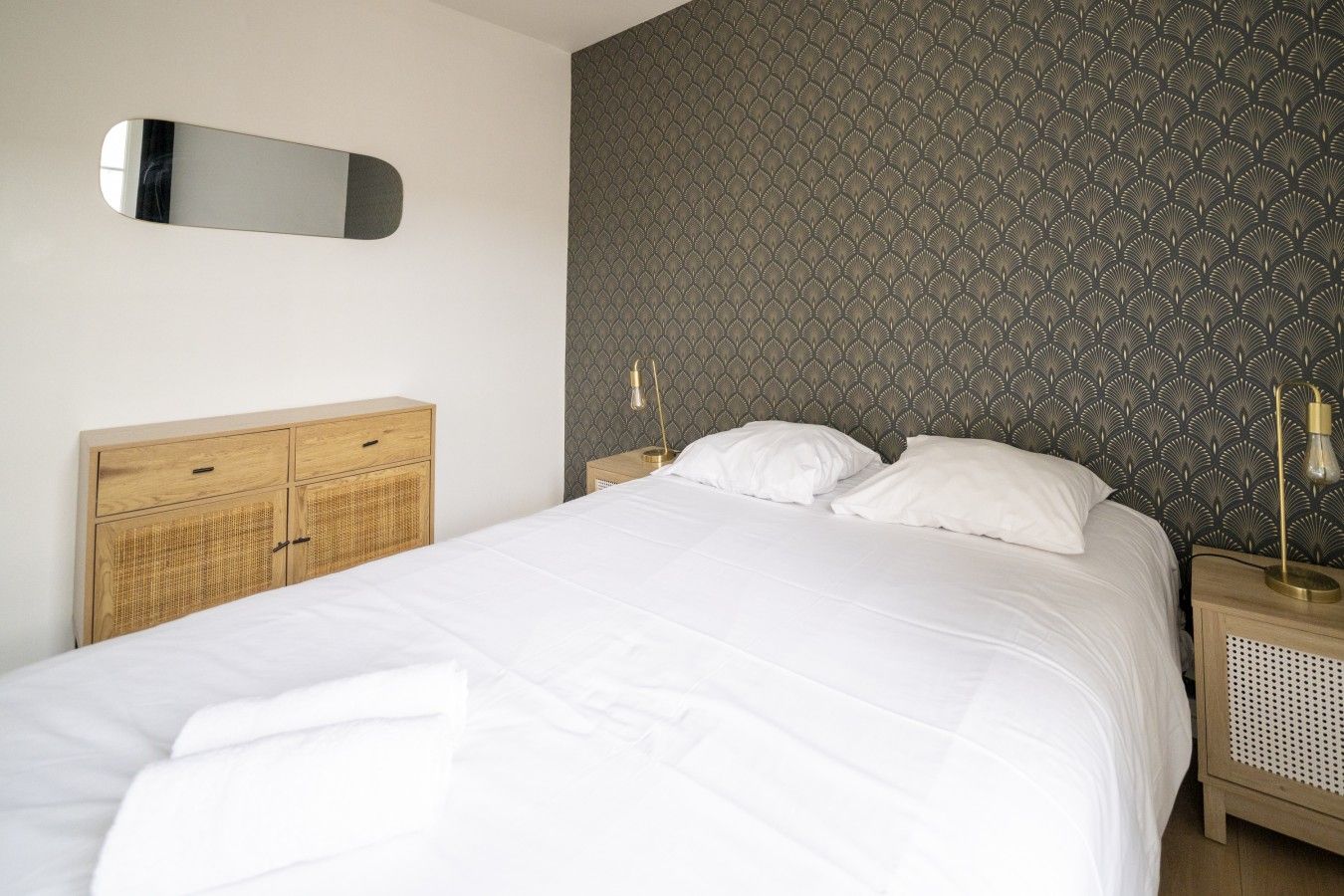 Un lit bien fait, avec du linge de lit blanc, une tête de lit en bois et un miroir fixé au mur.