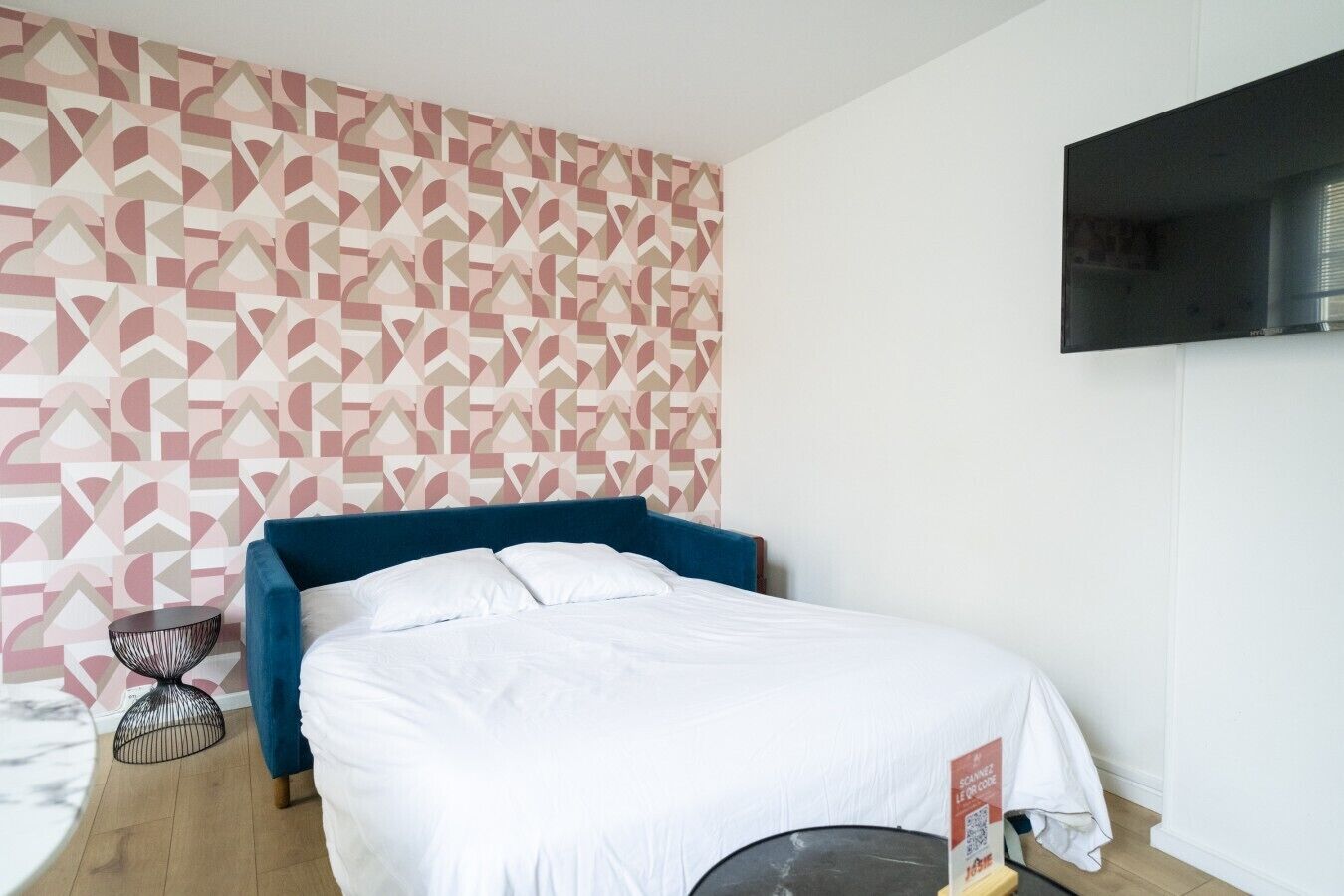 Een slaapkamer met een bed, een aan de muur bevestigde televisie, behang met een patroon en een klein nachtkastje.