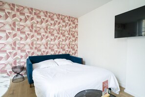 Een slaapkamer met een bed, een aan de muur bevestigde televisie, behang met een patroon en een klein nachtkastje.