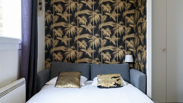 A bed with two pillows, a wall with a palm leaf pattern, a window with curtains, and a door. - Nice studio apartment for two, near the university (Caen)