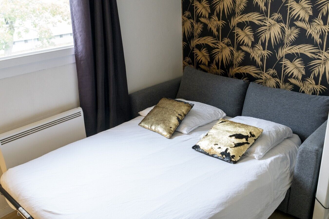 Una cama con ropa blanca y dos almohadas de diseño dorado y negro, un cabecero gris y una ventana con cortina.